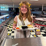 Ellieana joined the competition — help win amazing prizes! child, cafeteria, lunch, tray, food, carrots, bread, milk, chocolate_milk, golden_crown, costume, toga, smile, person, indoor, table, bench, floor, window, lighting