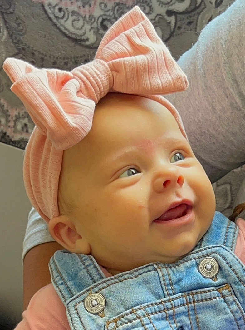 Kaylee joined the competition — help win amazing prizes! apparel, baby, background_pattern, child, closeup, cute, denim_overalls, face, fashion, happy, headband, holding, indoors, infant, person, pink_bow, portrait, skin, smiling, soft_lighting