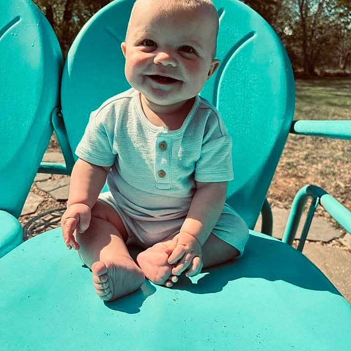 Kolter is registered to the contest to win money with this photo: baby, child, smiling, outdoor, chair, blue_chair, sunlight, grass, trees, happy, sitting, infant, casual_clothing, cute, one_person, daylight, nature, portrait, footwearless, young_child