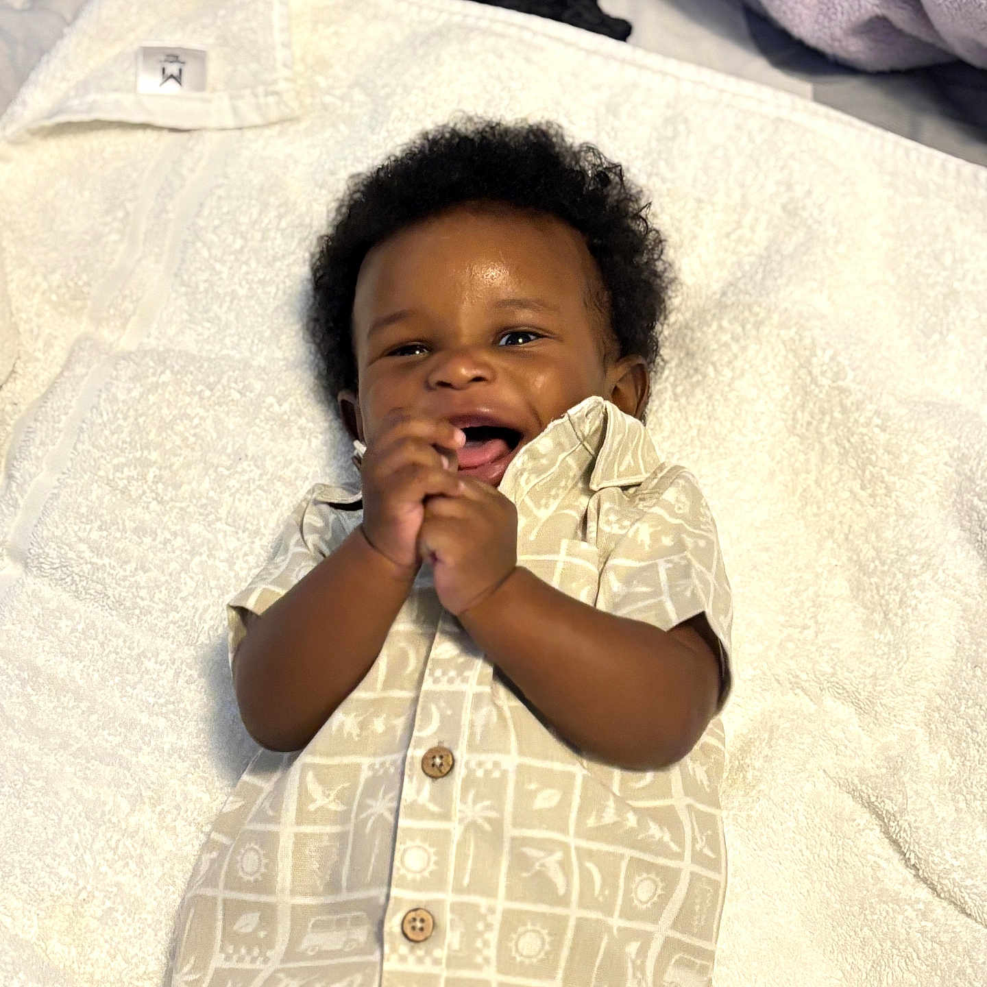 Remy is registered to the contest to win money with this photo: baby, child, smiling, clapping, towel, shirt, patterned_clothing, happy, infant, cute, lying_down, indoor, skin, curly_hair, portrait, small_hands, buttoned_shirt, comfort, soft_texture, light
