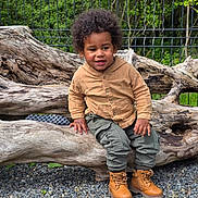 Amadou participe au concours pour gagner de l'argent avec cette photo : toddler, child, curly_hair, outdoor, bench, wood, gravel, greenery, shirt, pants, boots, smiling, nature, park, fence, happy, casual_clothing, portrait, sitting, daylight