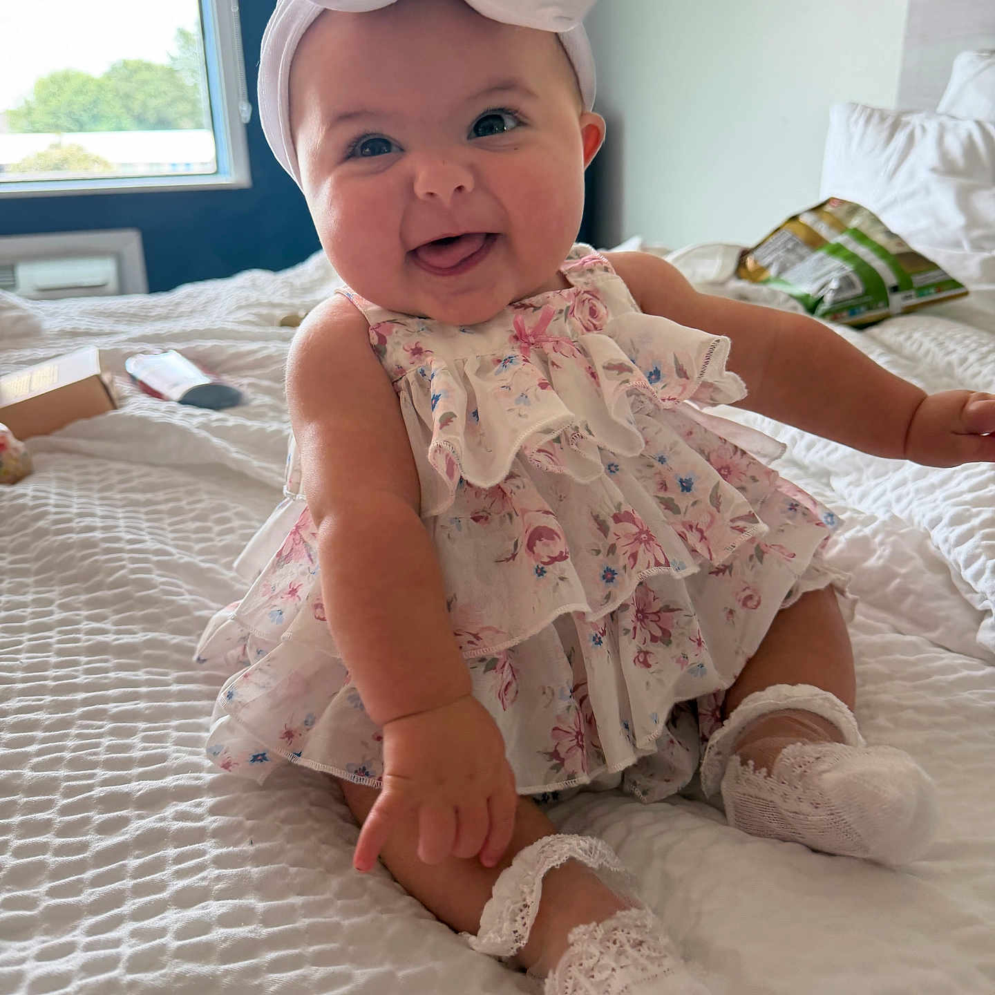 Hallee joined the competition — help win amazing prizes! baby, child, smiling, headband, bow, dress, floral, sitting, bed, white_bedspread, sock, cute, indoors, natural_light, happy, portrait, infant, room, cozy, window