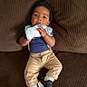 baby, child, infant, couch, brown, cloth, sitting, sneakers, suspenders, pants, blue_shirt, cute, indoors, portrait, face, hand, footwear, comfortable, young, fashion