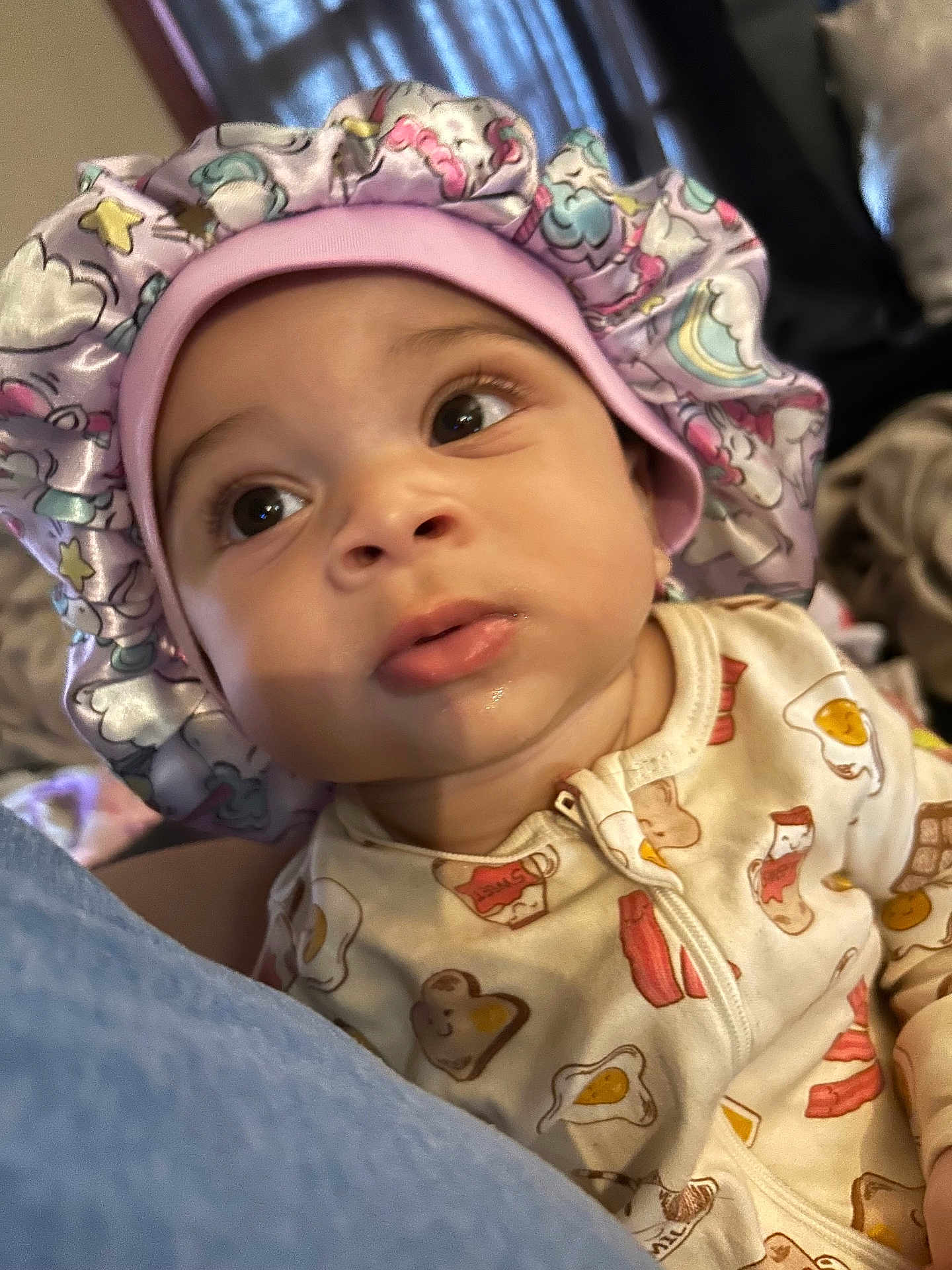 Zaierra is registered to the contest to win money with this photo: baby, infant, face, bonnet, hat, clothing, onesie, cute, expression, portrait, indoor, person, child, sleepwear, bedroom, soft_lighting, curious, unicorn_pattern, food_pattern, closeup