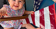 Andrea joined the competition — help win amazing prizes! baby, child, smiling, stroller, floral_dress, blue_wall, banner, red_stripes, white_stripes, happy, cute, person, infant, outdoor, hand, foot, face, portrait, seat, colorful