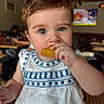 Andrea is registered to the contest to win money with this photo: baby, child, blue_eyes, white_dress, embroidered_clothing, eating, snack, indoor, restaurant, table, person, portrait, cute, young_child, face, hand, food, seated, casual, background_blur