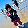 toddler, child, beach, sand, ocean, waves, hooded_jacket, maroon_pants, white_shoes, smiling, playing, sunny, outdoor, water, reflection, people, casual_clothing, daytime, nature, fun