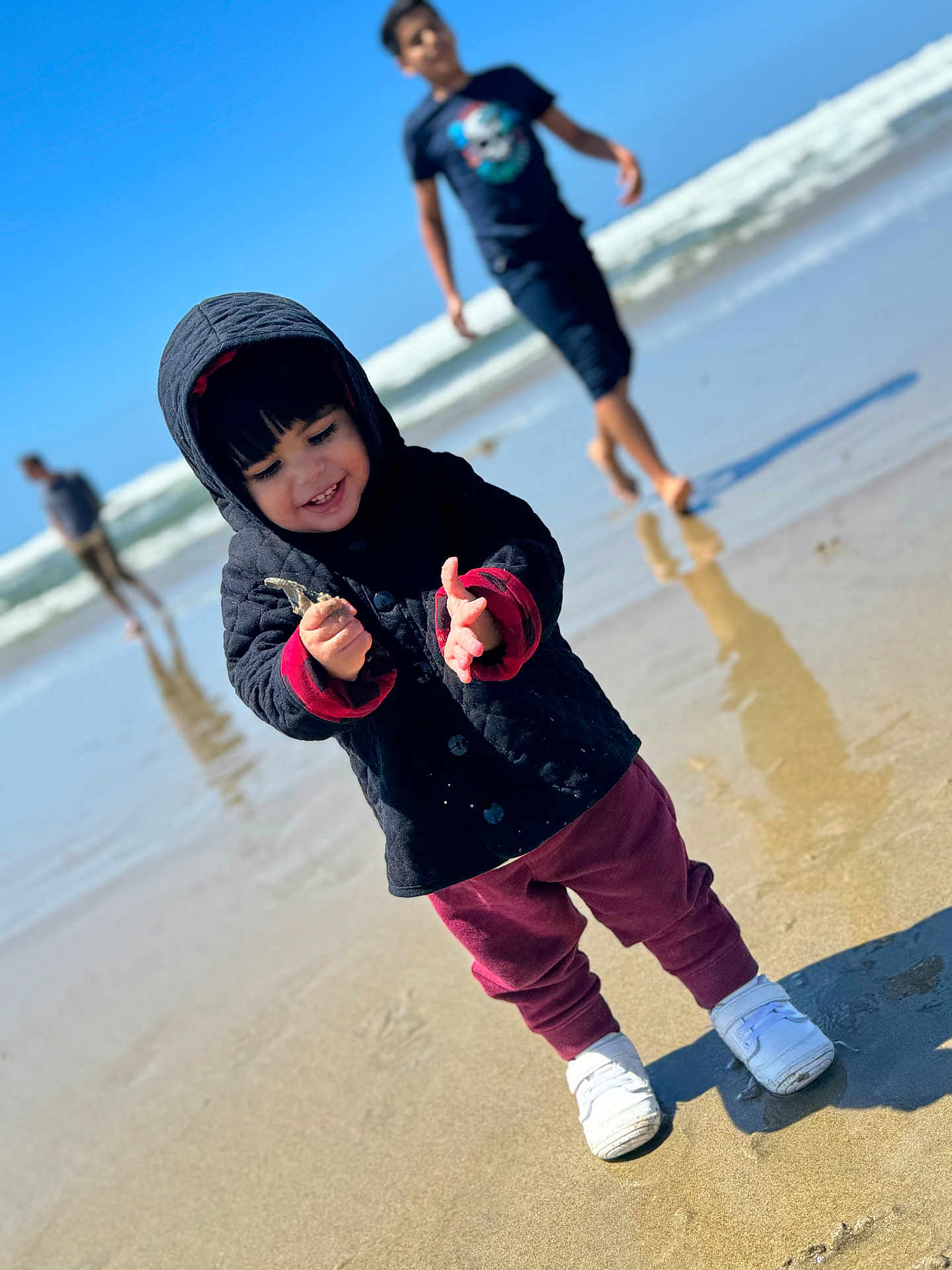 Billie is registered to the contest to win money with this photo: toddler, child, beach, sand, ocean, waves, hooded_jacket, maroon_pants, white_shoes, smiling, playing, sunny, outdoor, water, reflection, people, casual_clothing, daytime, nature, fun
