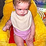 baby, child, bib, yellow_chair, pink_clothing, plush_chair, indoor, face, expression, person, sitting, floor_mat, toy, fan, home, rainbow_light, legs, hand, head, couch