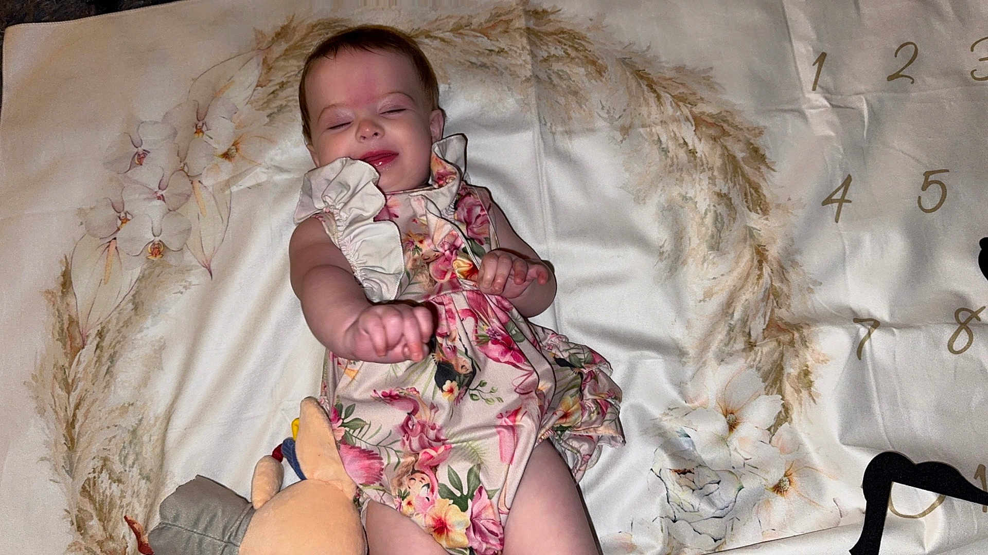Brynlee is registered to the contest to win money with this photo: baby, child, floral_dress, plush_toy, blanket, flower_pattern, numbers, smiling, closed_eyes, infant, cute, lying_down, soft_toy, fabric, home, portrait, person, baby_clothing, happy, infancy