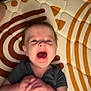 baby, infant, child, mat, pattern, clothing, hands, mouth_open, lying_down, face, expression, indoor, blur, skin, hair, person, young_child, play_area, colorful, candid