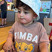 Fatir Hussain Mahar is registered to the contest to win money with this photo: toddler, child, hat, bucket_hat, simba_tshirt, high_chair, restaurant, indoor, table, person, casual_clothing, brown_shirt, shorts, seated, face, hands, expression, light, background, adult