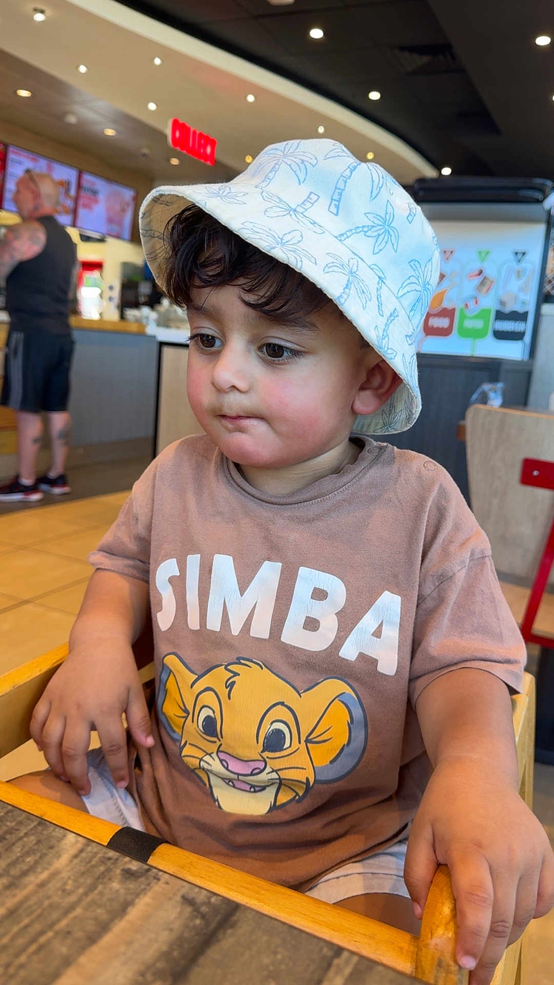 Fatir Hussain Mahar is registered to the contest to win money with this photo: toddler, child, hat, bucket_hat, simba_tshirt, high_chair, restaurant, indoor, table, person, casual_clothing, brown_shirt, shorts, seated, face, hands, expression, light, background, adult