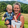 Asher is registered to the contest to win money with this photo: woman, toddler, child, overalls, blueberries, bucket, outdoor, field, greenery, nature, sky, clouds, smile, happy, holding, dreadlocks, casual_clothing, summer, family, portrait