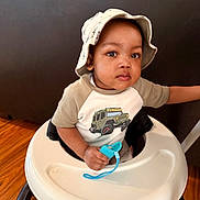 Imran joined the competition — help win amazing prizes! toddler, child, baby_walker, hat, bucket_hat, indoor, wooden_floor, pacifier, holding, shirt, jeep_print, furniture, couch, person, face, looking, cute, expression, sitting, young_child