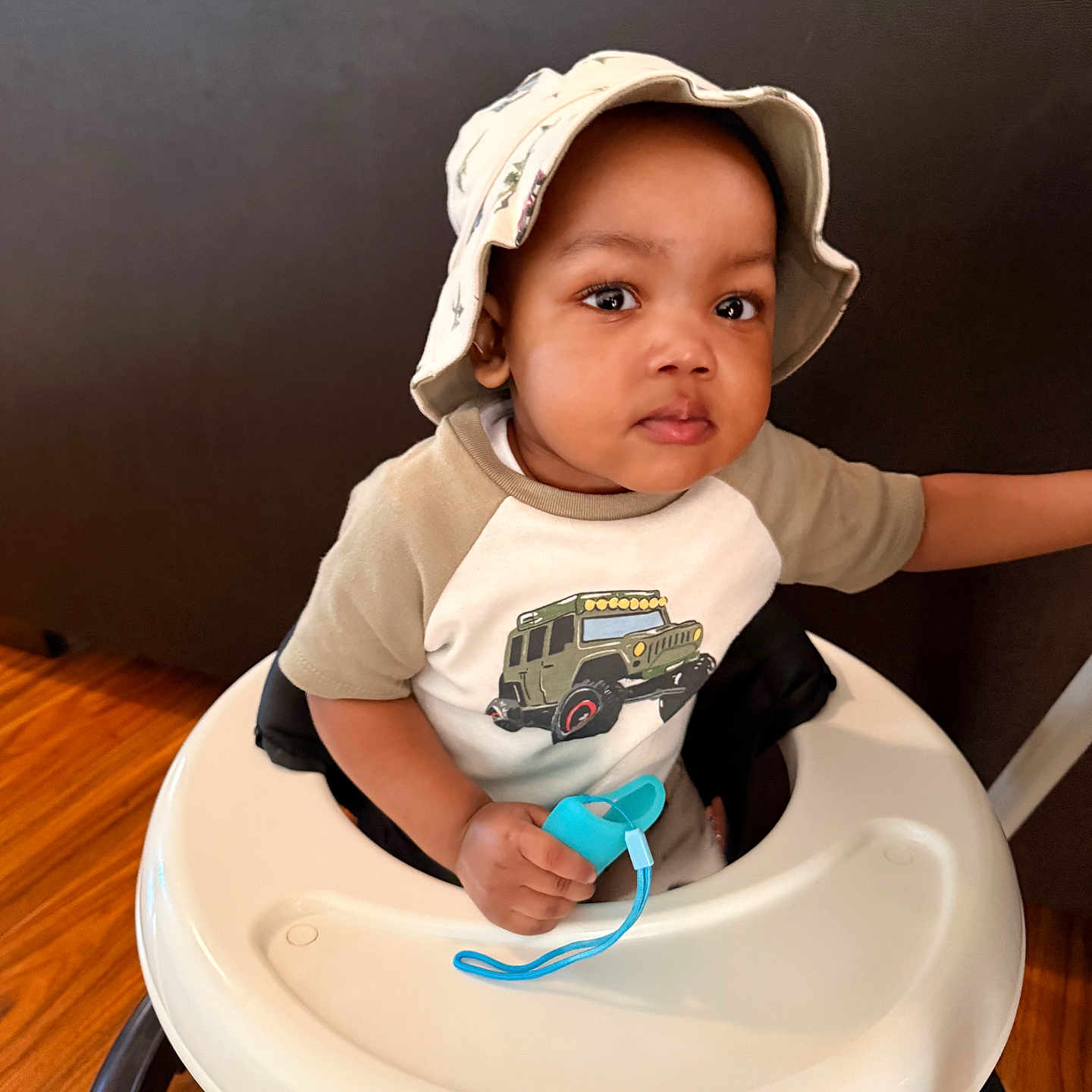 Imran joined the competition — help win amazing prizes! toddler, child, baby_walker, hat, bucket_hat, indoor, wooden_floor, pacifier, holding, shirt, jeep_print, furniture, couch, person, face, looking, cute, expression, sitting, young_child