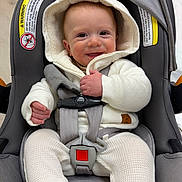 Santino is registered to the contest to win money with this photo: baby, infant, car_seat, smiling, white_clothing, hooded_jacket, straps, safety_buckle, cute, happy, child, indoors, warm_clothing, knit_pants, person, seat, infant_toddler, baby_face, closeup, portrait