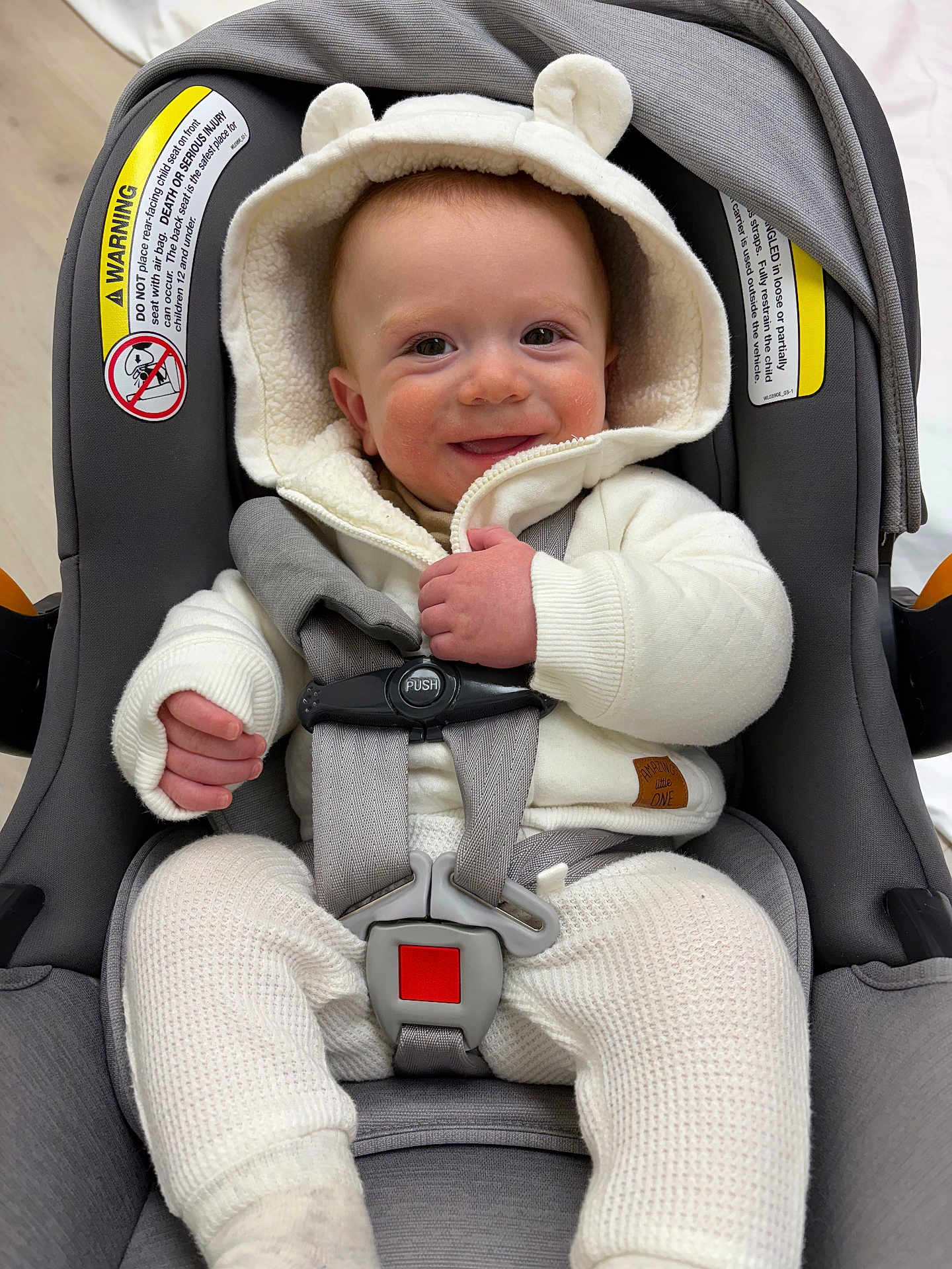 Santino is registered to the contest to win money with this photo: baby, infant, car_seat, smiling, white_clothing, hooded_jacket, straps, safety_buckle, cute, happy, child, indoors, warm_clothing, knit_pants, person, seat, infant_toddler, baby_face, closeup, portrait