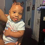 A'Lani is registered to the contest to win money with this photo: baby, bedroom, cartoon_shirt, child, cute, eyes, face, family, hair_buns, held, home, indoor, infant, person, portrait, sitting, smiling, striped_clothing, television, top_knots