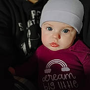 Olive joined the competition — help win amazing prizes! baby, child, beanie, clothing, face, blue_eyes, glossy_lips, portrait, indoor, person, hoodie, dark_background, close_up, cute, infant, apparel, text_on_clothing, rainbow, looking_at_camera, soft_lighting