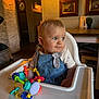 baby, high_chair, toy, indoor, child, blue_eyes, denim_dress, curly_hair, table, wall_art, wooden_frame, lighting, furniture, person, portrait, cute, infant, room, decor, play
