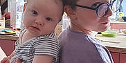 Oscar participe au concours pour gagner de l'argent avec cette photo : baby, child, sibling, glasses, striped_clothing, purple_shirt, kitchen, refrigerator, countertop, cup, water_bottle, cabinet, face, portrait, indoor, person, holding, smile, hair, hand