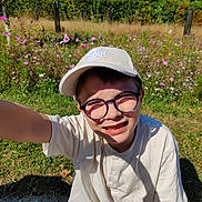 Arthur a rejoint le concours — aidez-le/la à gagner de superbes lots ! child, boy, glasses, cap, white_shirt, outdoor, sunlight, smiling, flower_garden, greenery, nature, daylight, grass, casual_clothing, portrait, young, happy, sitting, closeup, summer