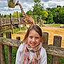child, girl, scarf, smile, fence, giraffe, animal, zoo, outdoor, grass, tree, cloudy_sky, wood, nature, portrait, happy, daylight, casual_clothing, young, cute