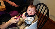 Eric joined the competition — help win amazing prizes! baby, high_chair, feeding, spoon, indoor, child, person, wooden_chair, table, hand, food, clothing, bib, floor, wall, sitting, expression, face, adult, casual