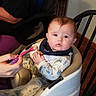 baby, high_chair, feeding, spoon, indoor, child, person, wooden_chair, table, hand, food, clothing, bib, floor, wall, sitting, expression, face, adult, casual