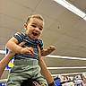 child, toddler, smiling, happy, person, hands, indoor, store, shopping, appliances, fluorescent_lighting, ceiling, clothing, striped_shirt, green_pants, hair_accessory, lifting, joy, playful, retail