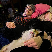 Monty joined the competition — help win amazing prizes! man, glasses, beanie, mustache, cat, bed, pillow, hand, black_clothing, tattoo, phone, indoor, relaxed, lying_down, feline, pet, cozy, cuddling, pink_pillow, casual