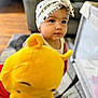 toddler, child, headband, plush_toy, winnie_the_pooh, indoor, living_room, carpet, couch, playpen, curly_hair, white_clothing, red_clothing, toy, baby, person, cute, soft_toy, expression, gaze