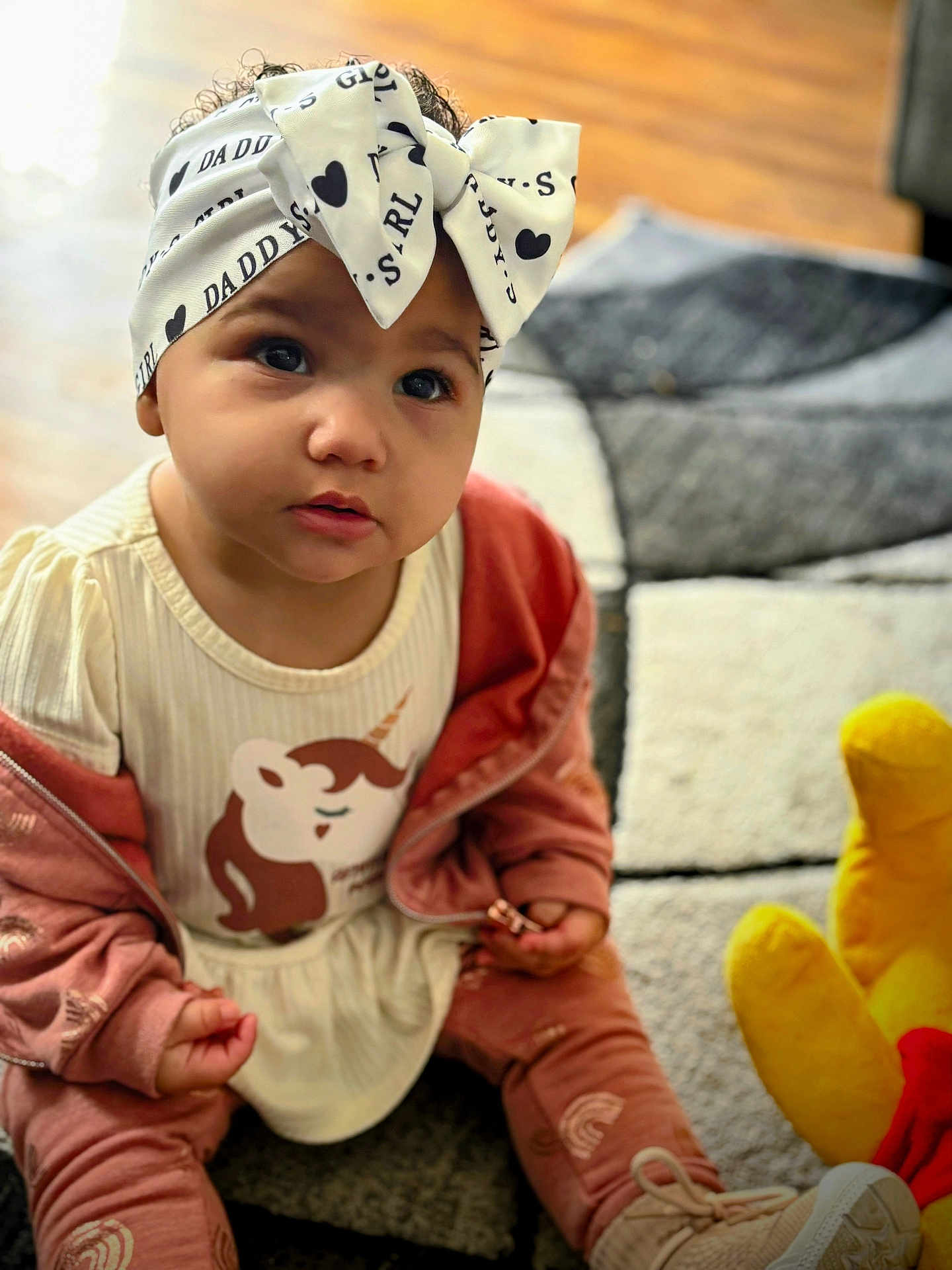 Isabelle is registered to the contest to win money with this photo: toddler, child, headband, bow, pink_clothing, sitting, rug, indoor, toy, yellow_plush, curious, face, expression, hands, footwear, patterned, floor, young_child, portrait, casual
