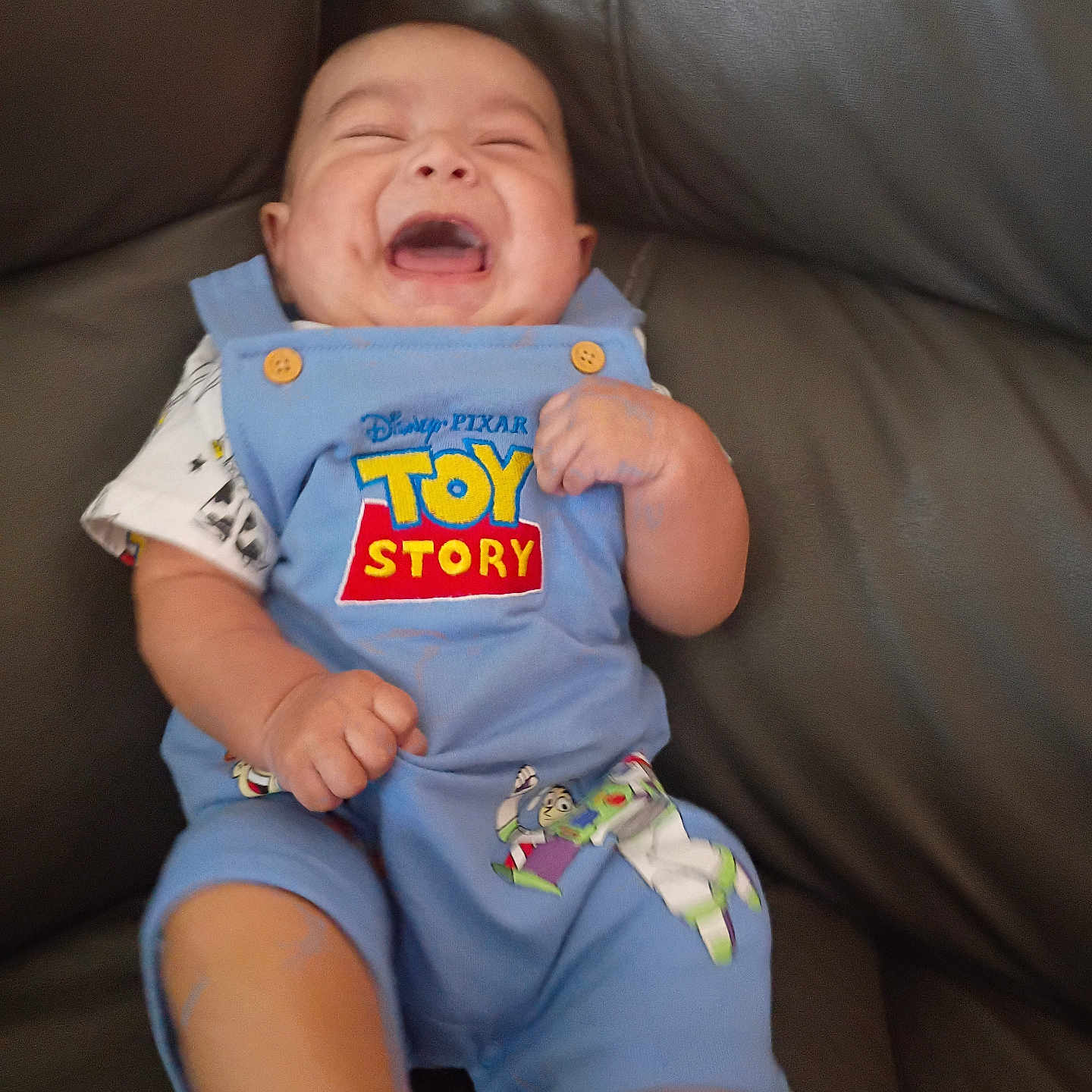 Robert is registered to the contest to win money with this photo: baby, blue_overall, button, child, clothing, couch, cute, fist, happy, indoor, infant, joyful, laughing, leather, person, playful, seated, short_sleeve, smiling, toy_story