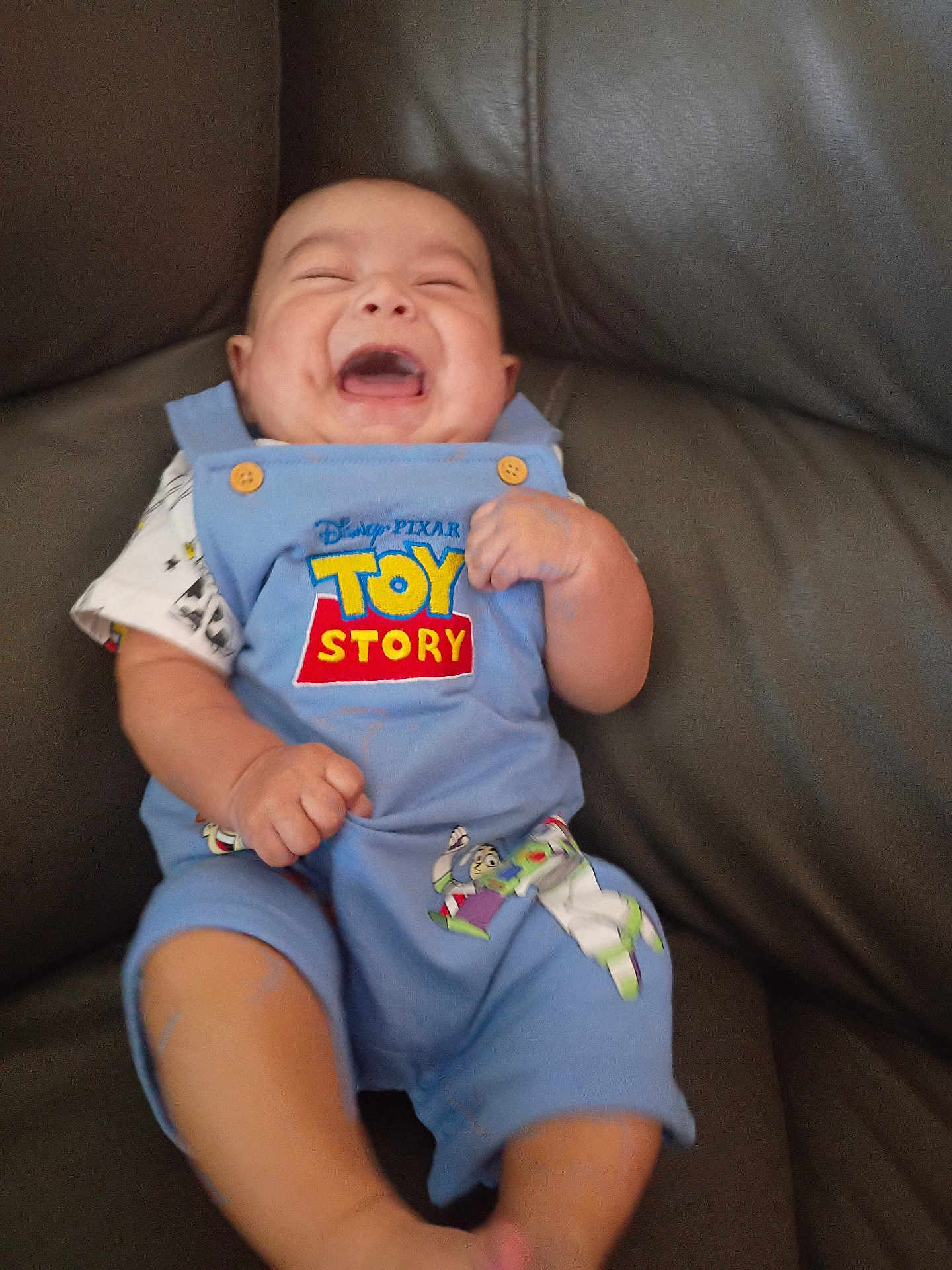 Robert is registered to the contest to win money with this photo: baby, child, laughing, toy_story, blue_overall, couch, leather, happy, infant, smiling, clothing, seated, cute, joyful, indoor, person, playful, short_sleeve, fist, button