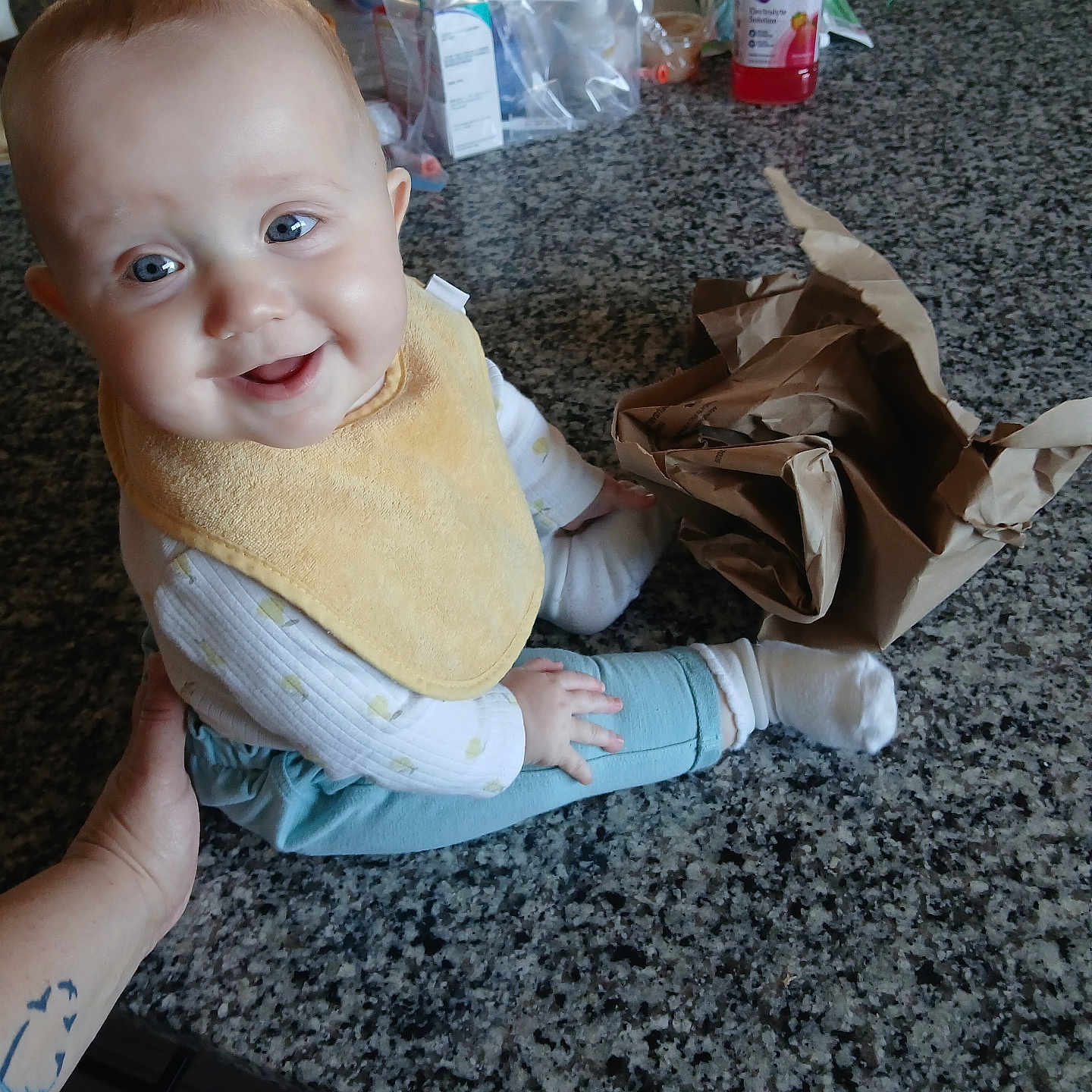 April joined the competition — help win amazing prizes! baby, child, infant, smiling, yellow_bib, blue_eyes, tattoo, hand, granite_countertop, kitchen, bottle, paper_bag, clothing, sitting, happy, person, indoor, table, food_container, curious