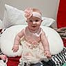 April is registered to the contest to win money with this photo: baby, child, smiling, pink_dress, headband, flower, cushion, bed, pillow, blanket, indoor, cute, happy, person, infant, toddler, clothing, face, sitting, home