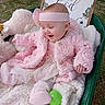 baby, pink_clothing, headband, blanket, wheelbarrow, toy, outdoor, grass, smiling, infant, cute, child, happy, portrait, soft_texture, decorative_sign, warm_clothing, playful, seated, daylight