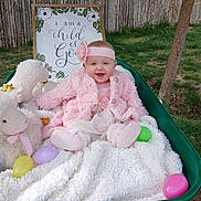 April is registered to the contest to win money with this photo: baby, child, pink_clothing, headband, wheelbarrow, blanket, stuffed_animal, plastic_eggs, outdoor, grass, tree, wooden_fence, sign, smiling, happy, portrait, cute, cozy, spring, playful