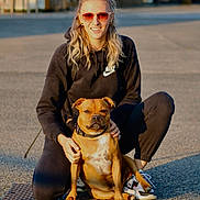 Tao a rejoint le concours — aidez-le/la à gagner de superbes lots ! woman, dog, outdoor, pavement, sunlight, sunglasses, black_hoodie, nike, kneeling, pet, brown_dog, casual_clothing, smiling, daylight, urban, leisure, friendly, bond, portrait, shoes
