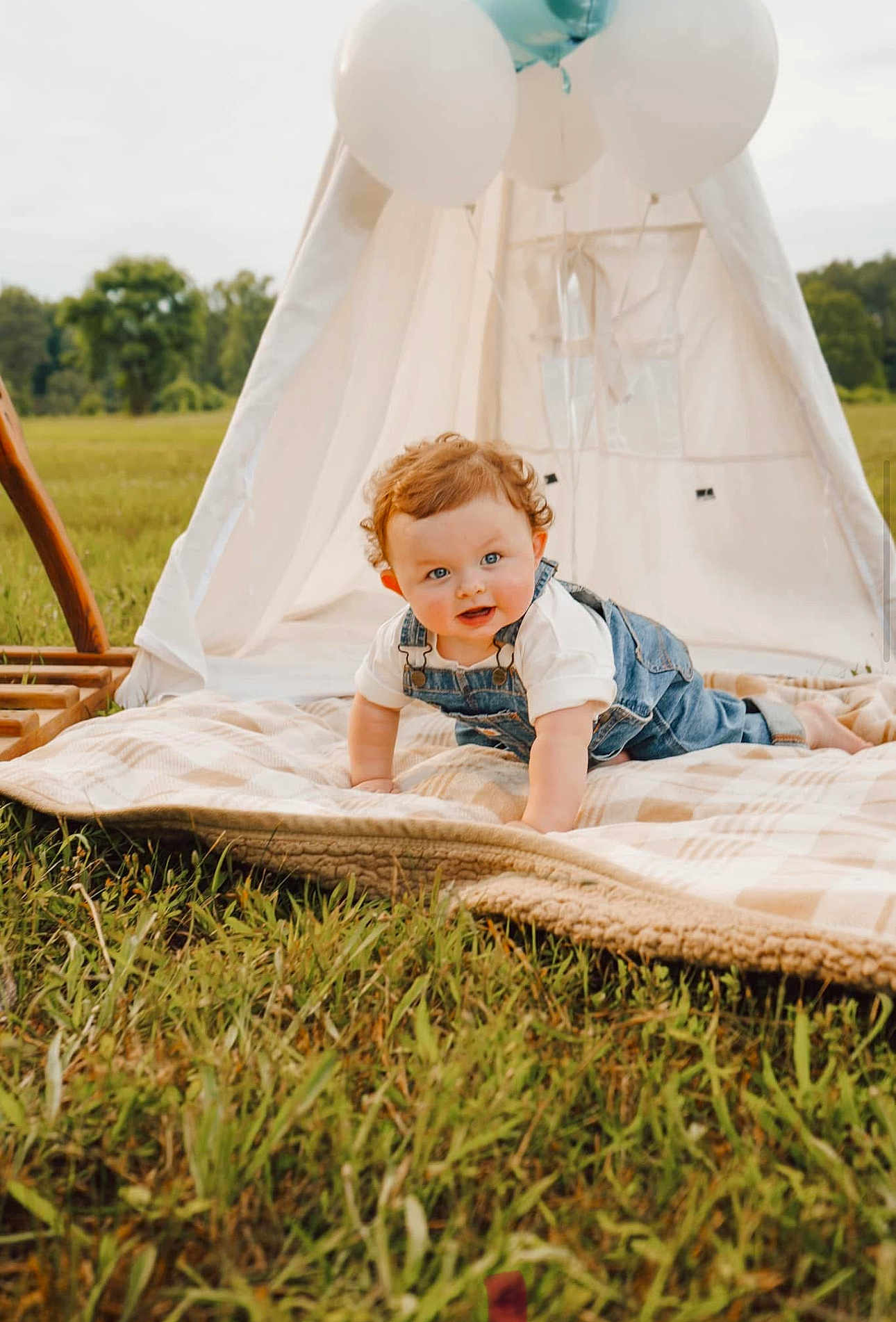 Walker is registered to the contest to win money with this photo: baby, toddler, infant, curly_hair, denim_overalls, blanket, teepee, balloons, grass, field, outdoor, portrait, crawling, smile, blue_eyes, picnic, playful, sunlight, nature, family