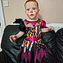 Willow is registered to the contest to win money with this photo: toddler, child, costume, face_paint, clown, dress, striped_tights, red_hair, indoor, couch, black_leather, wall, curious, standing, colorful, patterned, portrait, person, home, fun