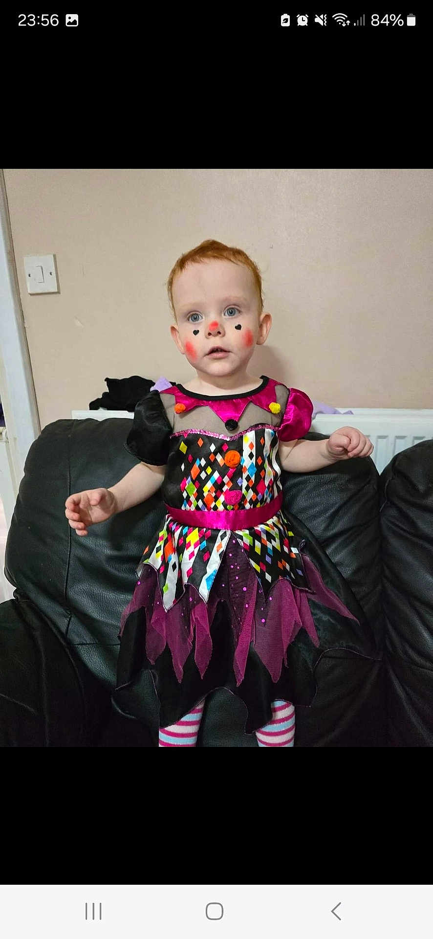 Willow is registered to the contest to win money with this photo: toddler, child, costume, face_paint, clown, dress, striped_tights, red_hair, indoor, couch, black_leather, wall, curious, standing, colorful, patterned, portrait, person, home, fun