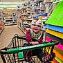 baby, infant, child, shopping_cart, store_aisle, smiling, cranial_helmet, bib, socks, hands, dollar_tree, shelves, craft_supplies, colorful_paper, retail, floor, portrait, seat, shopping, happy