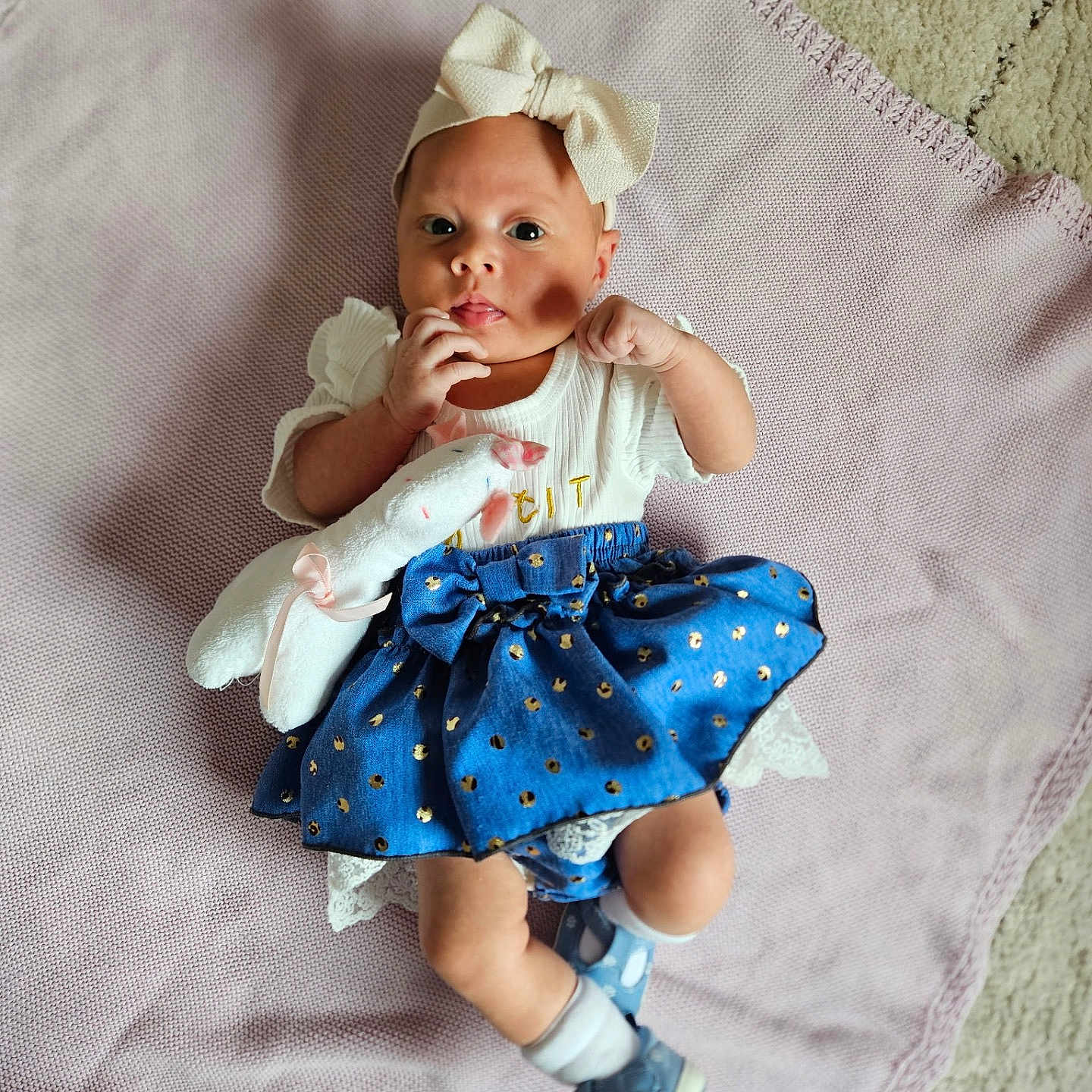 Arabella is registered to the contest to win money with this photo: baby, blanket, blue_skirt, bow, carpet, child, clothing, cute, floor, headband, indoors, infant, lying_down, person, plush_toy, portrait, shoes, socks, soft_toy, white_top