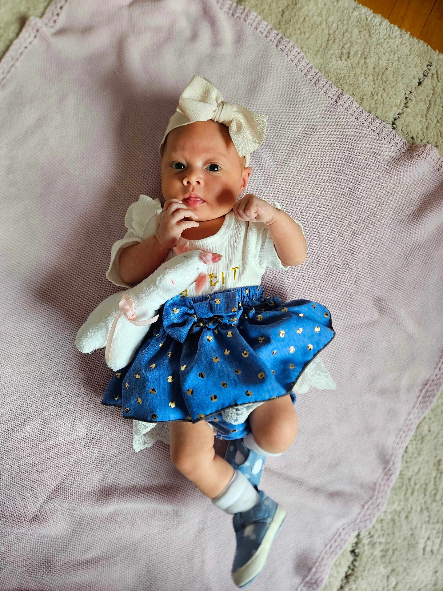 Arabella is registered to the contest to win money with this photo: baby, child, infant, blanket, plush_toy, headband, bow, blue_skirt, white_top, socks, shoes, floor, carpet, cute, lying_down, indoors, portrait, soft_toy, clothing, person