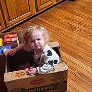 Athena is registered to the contest to win money with this photo: black_hearts, box, cabinets, cantaloupes, cardboard_box, child, curious, curly_hair, expression, flooring, food_packaging, grocery_box, indoor, kitchen, pantry_items, person, puzzled, toddler, white_shirt, wooden_floor