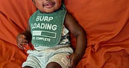 Dakota is registered to the contest to win money with this photo: baby, child, smiling, sitting, bib, clothing, socks, shorts, orange_couch, furniture, face, person, indoors, happy, cute, home, infant, expression, head, seated