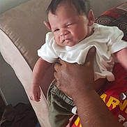 Jakai is registered to the contest to win money with this photo: baby, infant, person, hand, clothing, white_shirt, green_shorts, couch, furniture, indoor, expression, scrunched_face, holding, adult_hand, skin, casual, home, portrait, child, young